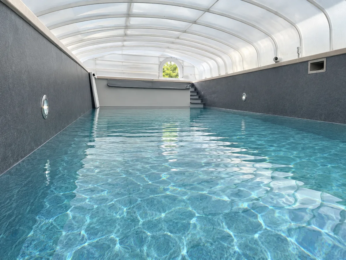 Piscine rectangulaire gris foncé sous abri cintré, eau claire, volet enroulé et margelles sombres
