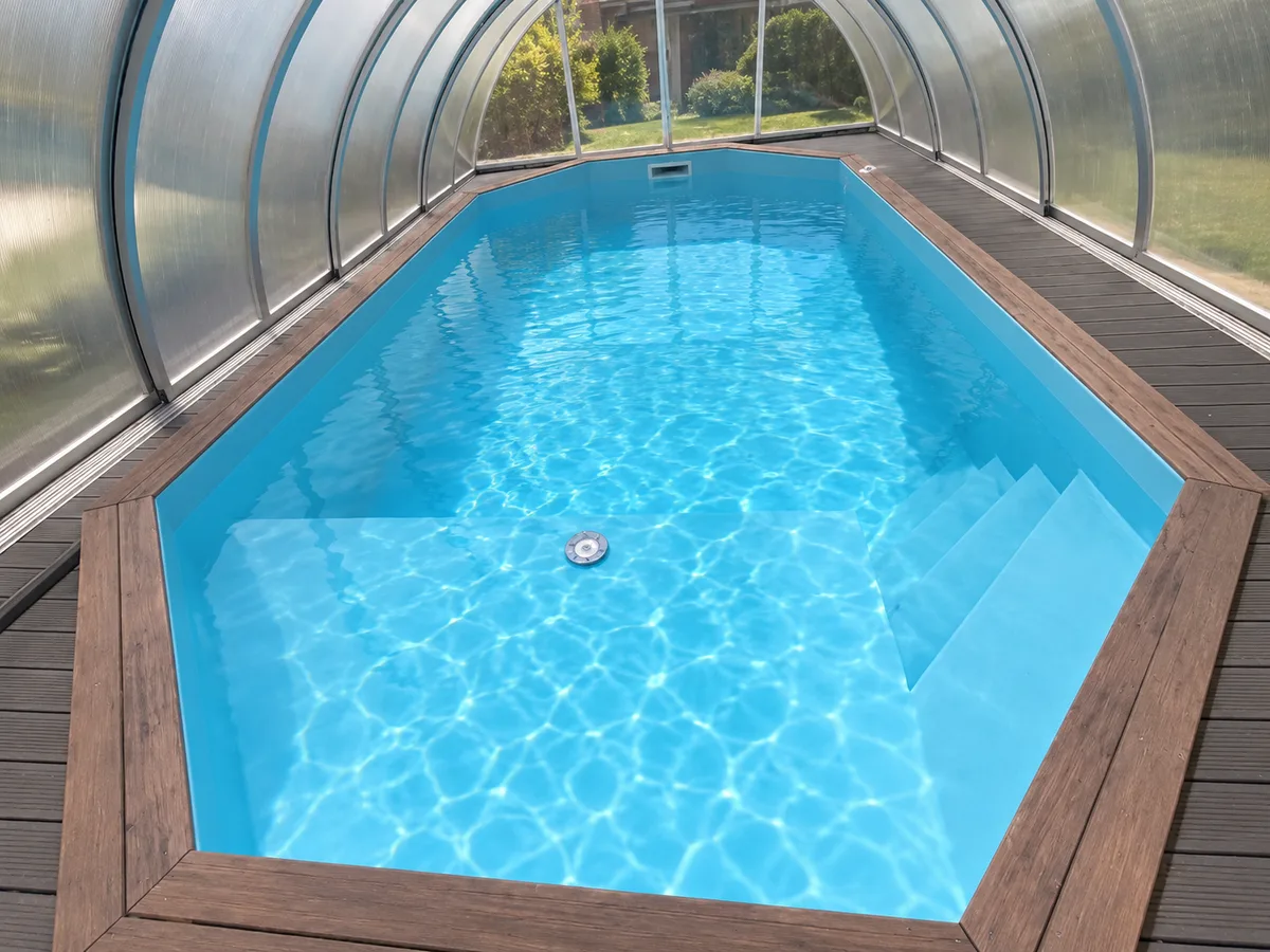 Bassin polygonal bleu clair sous abri transparent, eau turquoise, escalier immergé et terrasse en bois brun