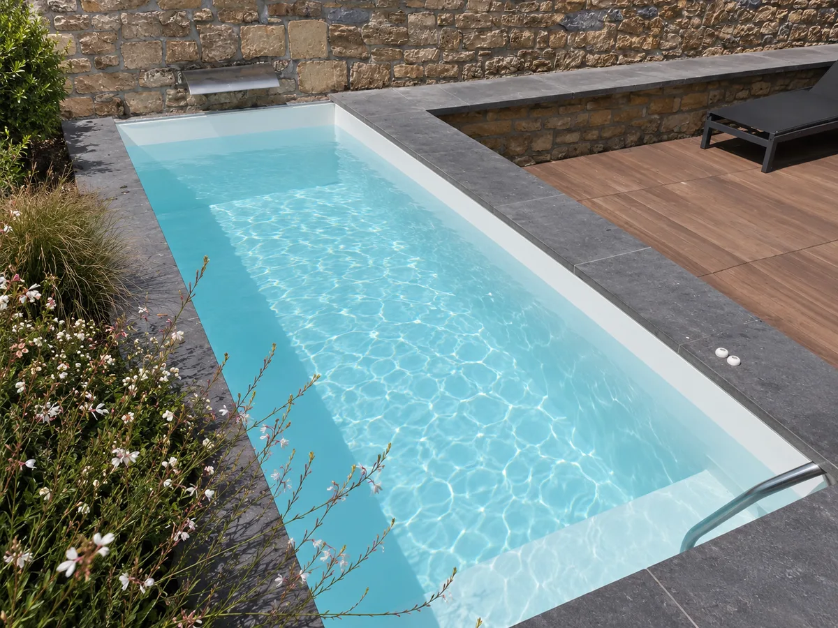 Bassin rectangulaire étroit gris clair, eau turquoise, margelles sombres, terrasse en bois, mur en pierre et massifs fleuris