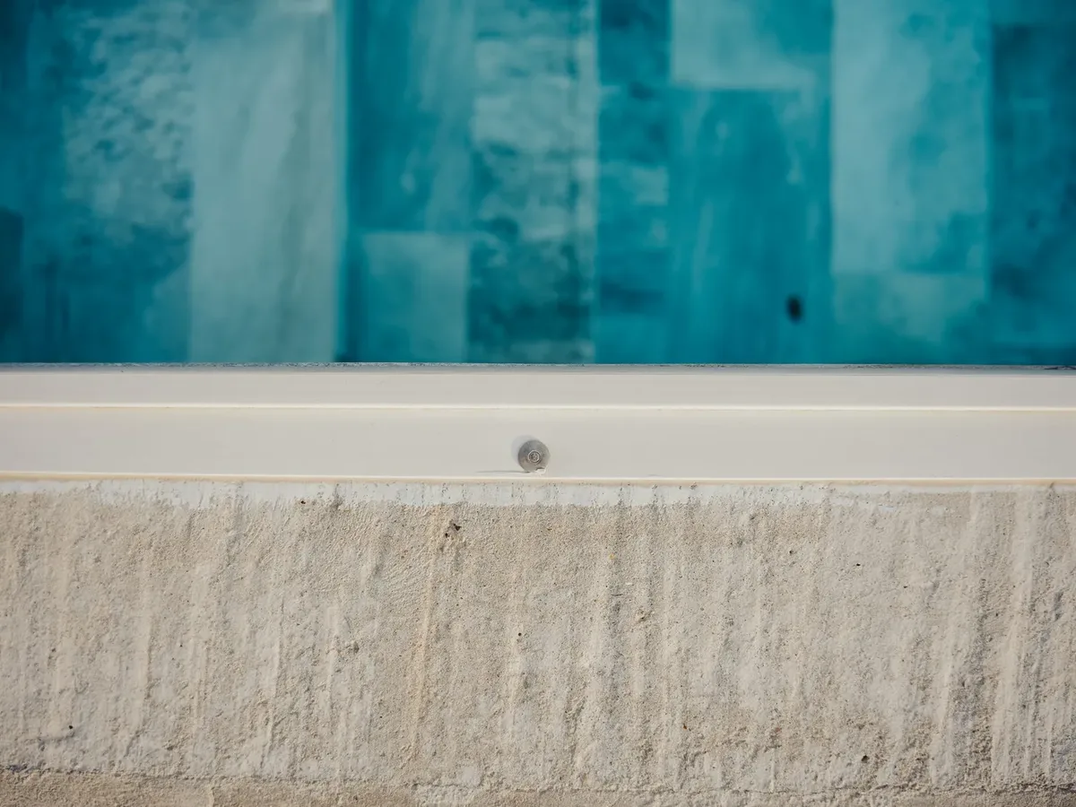 Détail du bord d'une piscine : bande claire sous l'eau avec un petit orifice métallique, maçonnerie brute au-dessus
