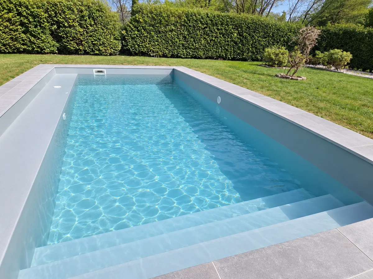 Bassin rectangulaire gris clair, eau turquoise, large escalier immergé, terrasse dallée grise, pelouse et haie