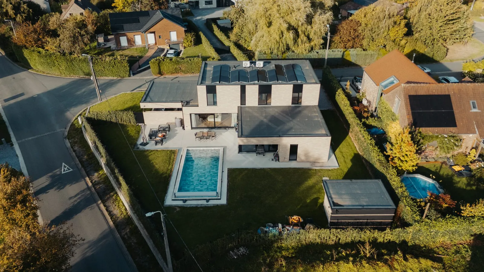 Vue aérienne d'une maison contemporaine en briques avec panneaux solaires sur le toit et sa piscine rectangulaire dans le jardin, au bord d'une route
