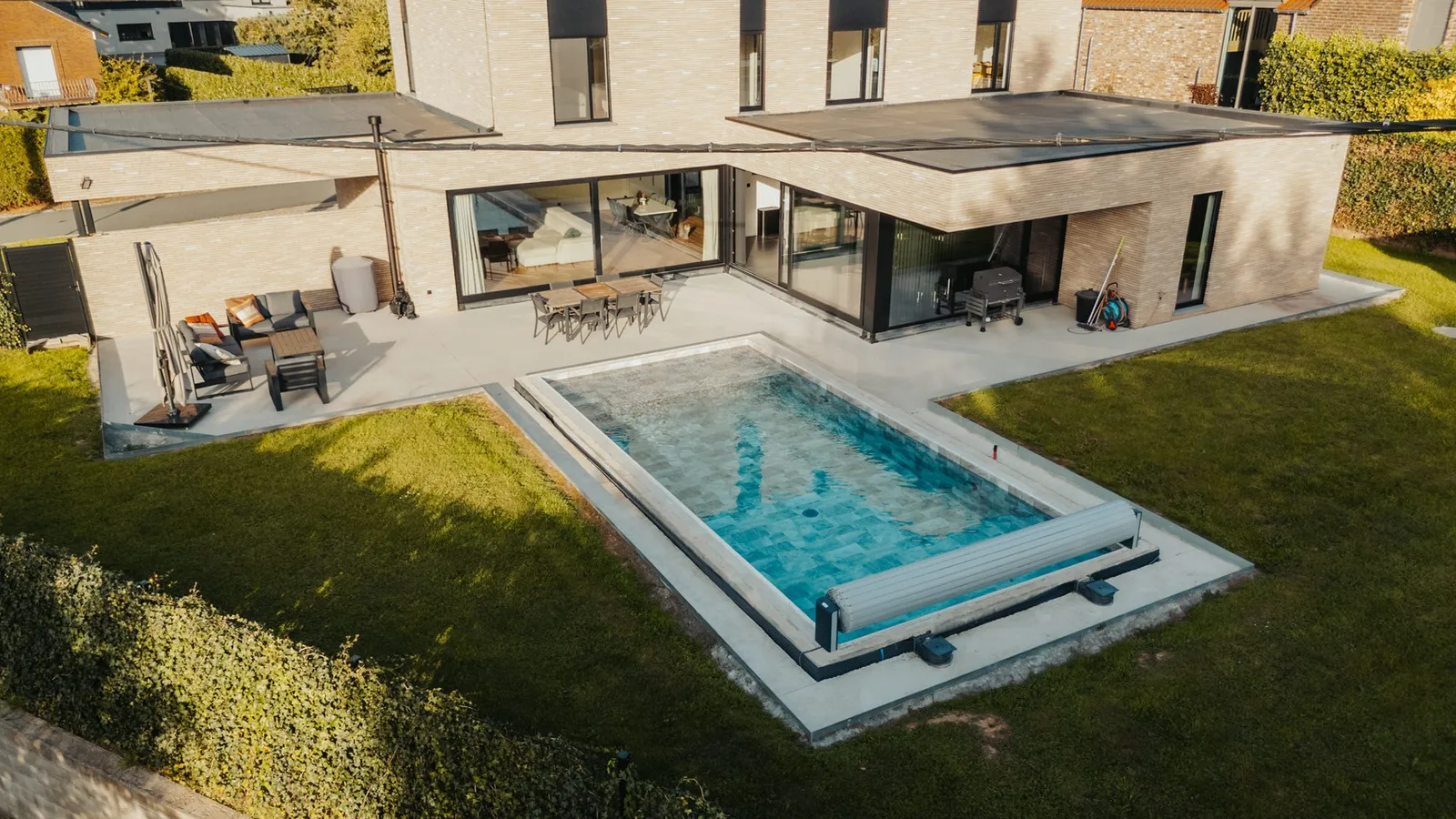 Vue aérienne rapprochée d'une piscine rectangulaire avec son volet roulant enroulé à une extrémité, terrasse et façade de la maison à l'arrière-plan