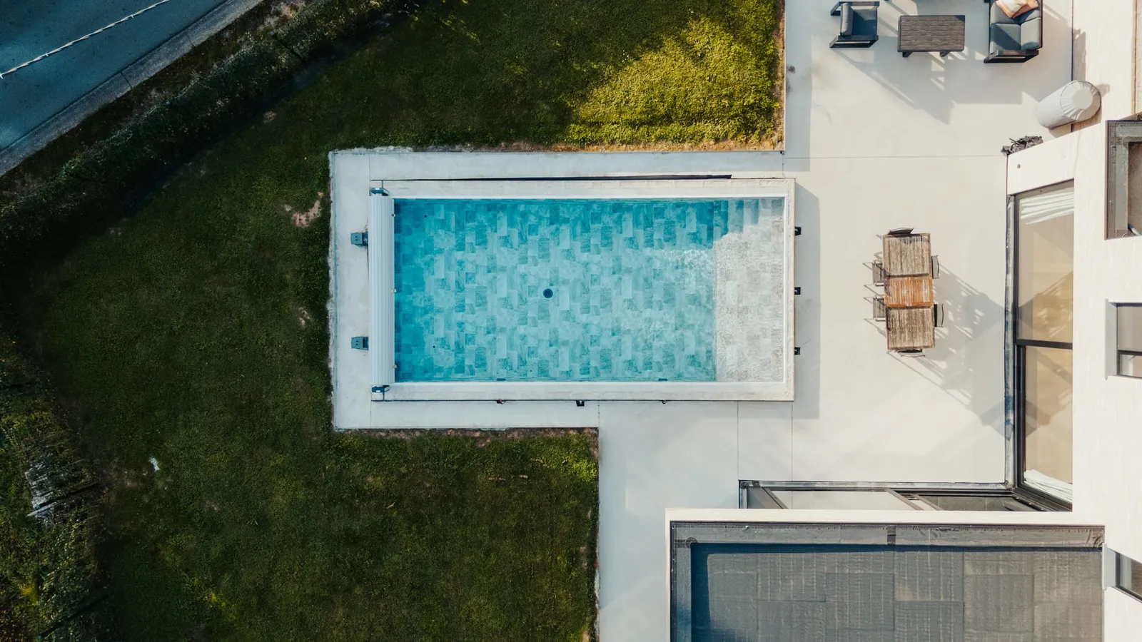Vue du dessus d'une piscine rectangulaire à fond bleu clair entourée d'une terrasse en béton clair, pelouse et mobilier de jardin sur le côté