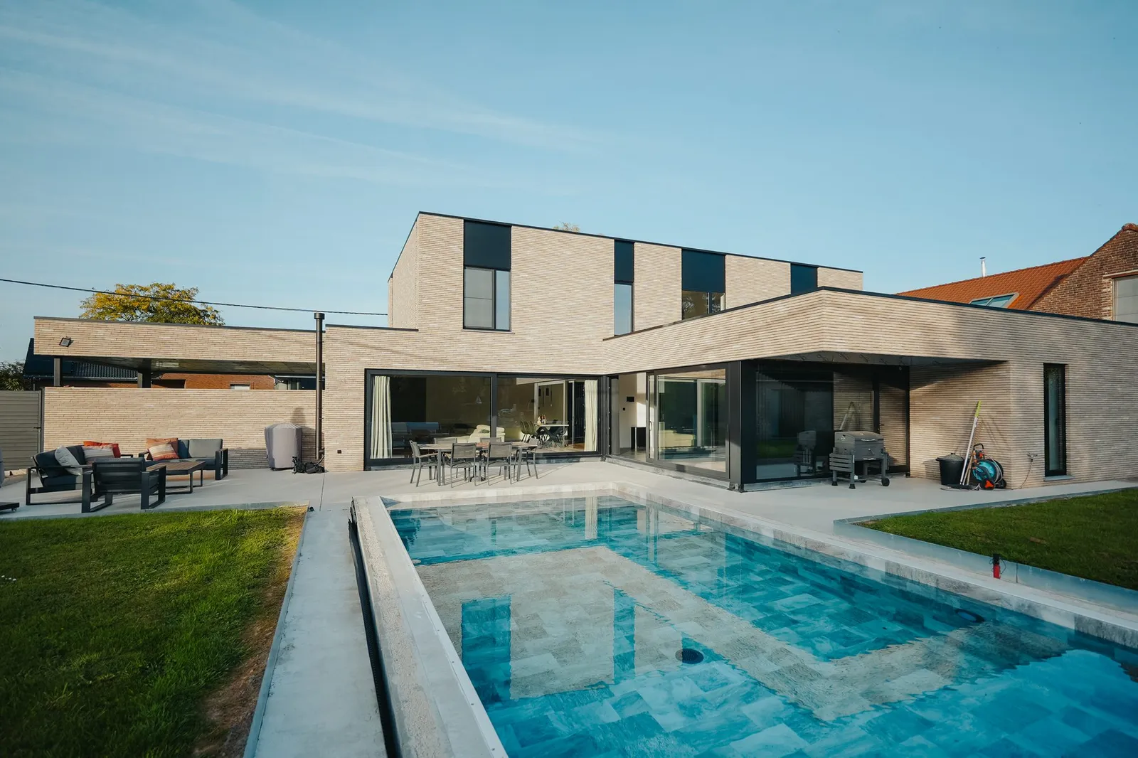Piscine longée par une fine rigole en béton, pelouse sur le côté et maison en briques avec grandes baies vitrées en fond
